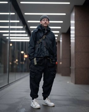 A next to a futuristic building in complete techwear outfit consisting of an ACRONYM techwear jacket, an ACRONYM techwear bag, ACRONYM P38-E pants and ACRONYM x Nike Lunar Force techwear sneaker