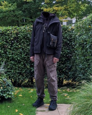 A person in standing in a garden in a techwear outfit with Adidas MyShelter techwear jacket, Enfin Levße Aurrean techwear pants and Hoka Tor Low techwear shoes and ACRONYM 3A-2 bag
